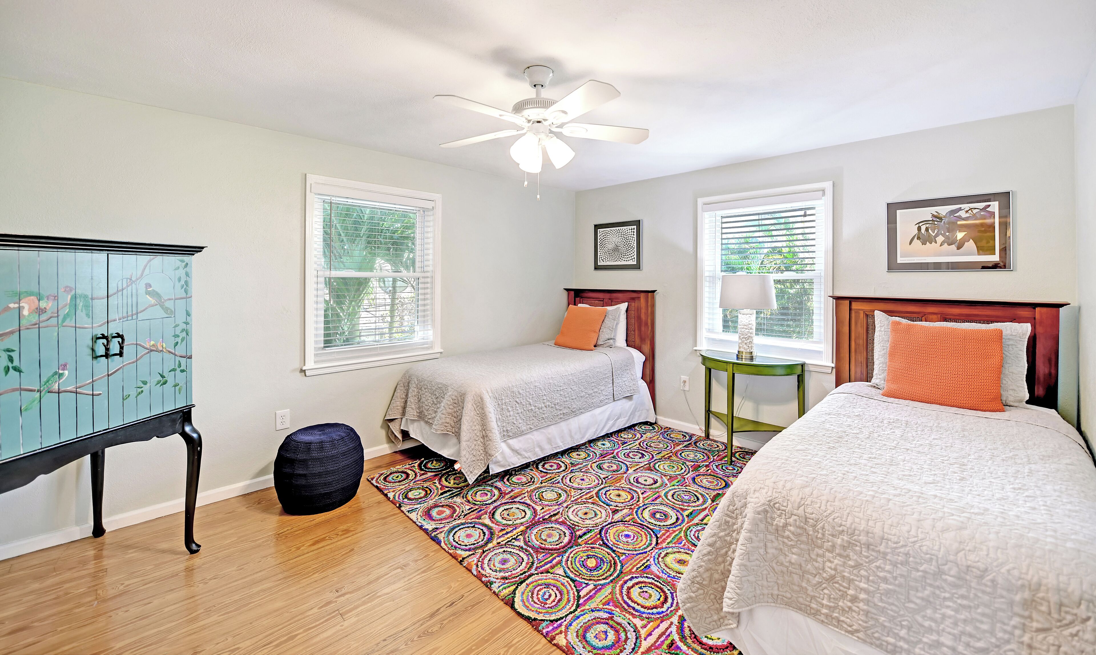 A bedroom with two beds, a ceiling fan, a window with blinds, a colorful rug, and a cabinet with a bird mural.