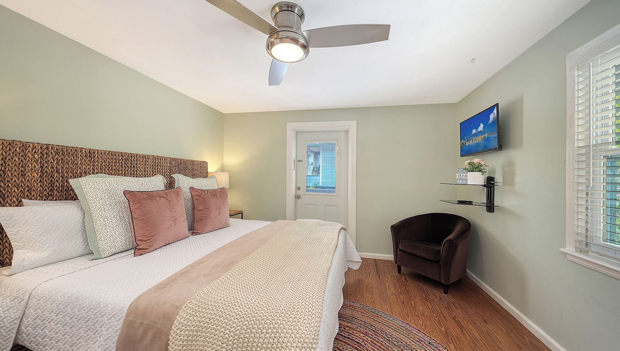A bedroom with a bed, a ceiling fan, a door with a window, a chair, a TV, and a small shelf.