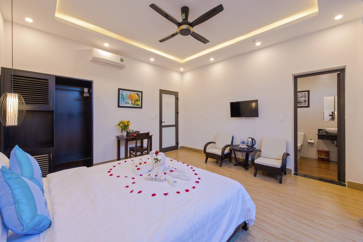 Quarto de hotel moderno com uma cama grande, ventoinha no teto, televisão, escrivaninha com cadeira e um pequeno espaço de estar.