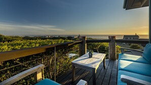 A wooden deck with a table and blue chairs, overlooking a beach with a clear sky. - DOGS, Panoramic Ocean Views, Game Room, EV Charger, New Hot Tub, Chef’s Kitchen (Waldport)