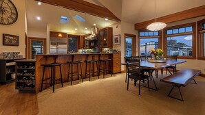 A spacious kitchen with a central island, bar stools, a dining table with chairs, and large windows offering views. - DOGS, Panoramic Ocean Views, Game Room, EV Charger, New Hot Tub, Chef’s Kitchen (Waldport)