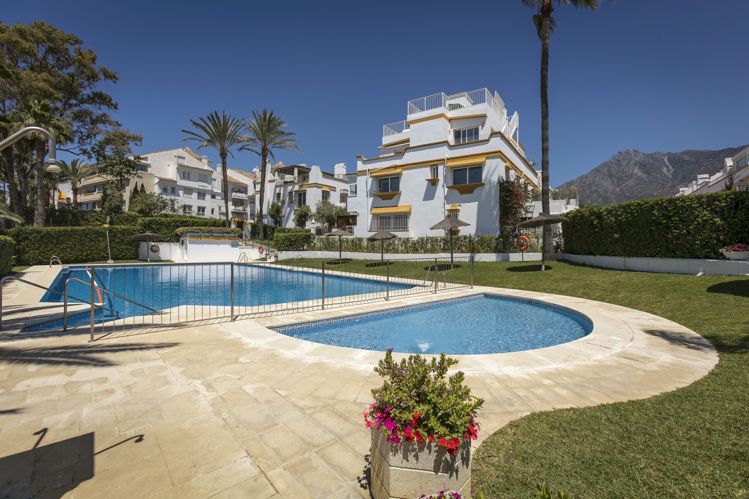 A swimming pool with a curved edge, surrounded by a paved deck. In the background, there are multi-story buildings with balconies and a mountain in the distance.