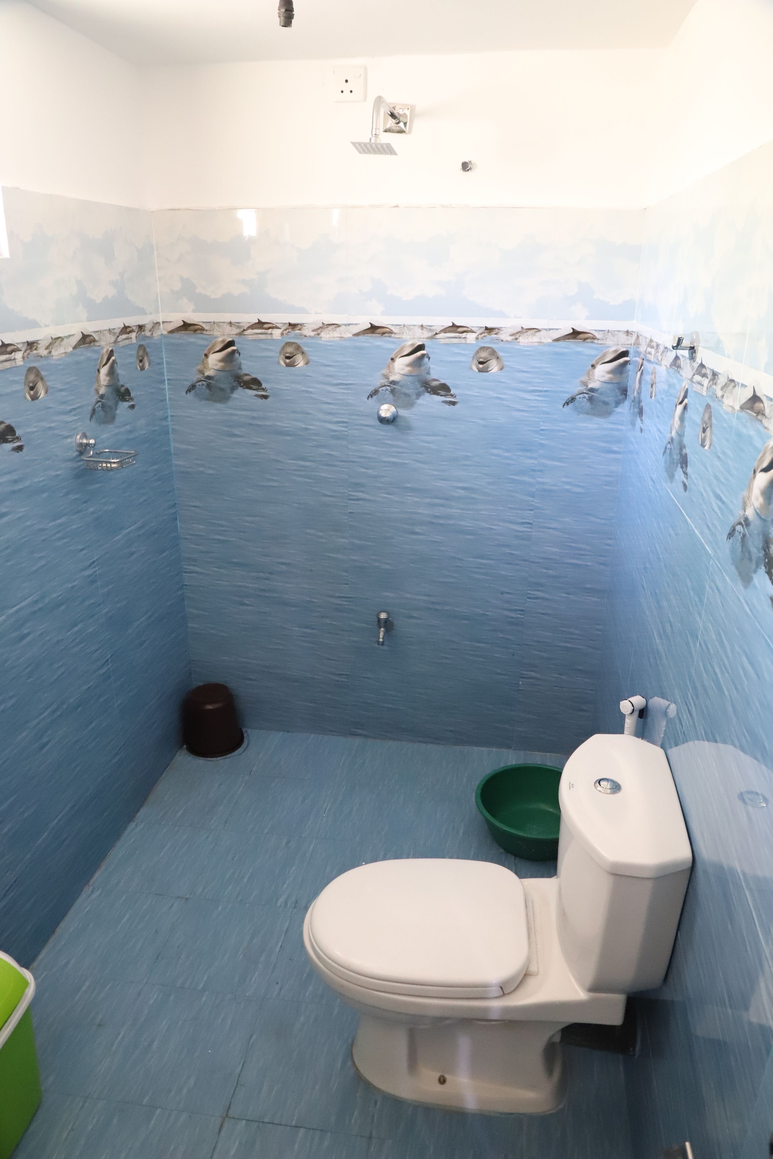 A bathroom with a blue tiled wall featuring dolphin murals, a white toilet, a green basin, and a brown trash bin.