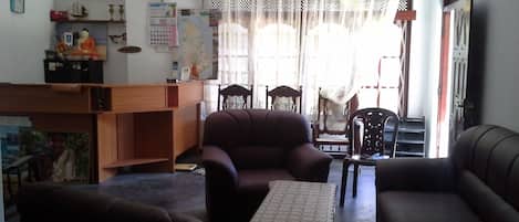 A living room with a sofa, armchair, and a small table. There is a wooden desk with a chair and a shelf with various items.