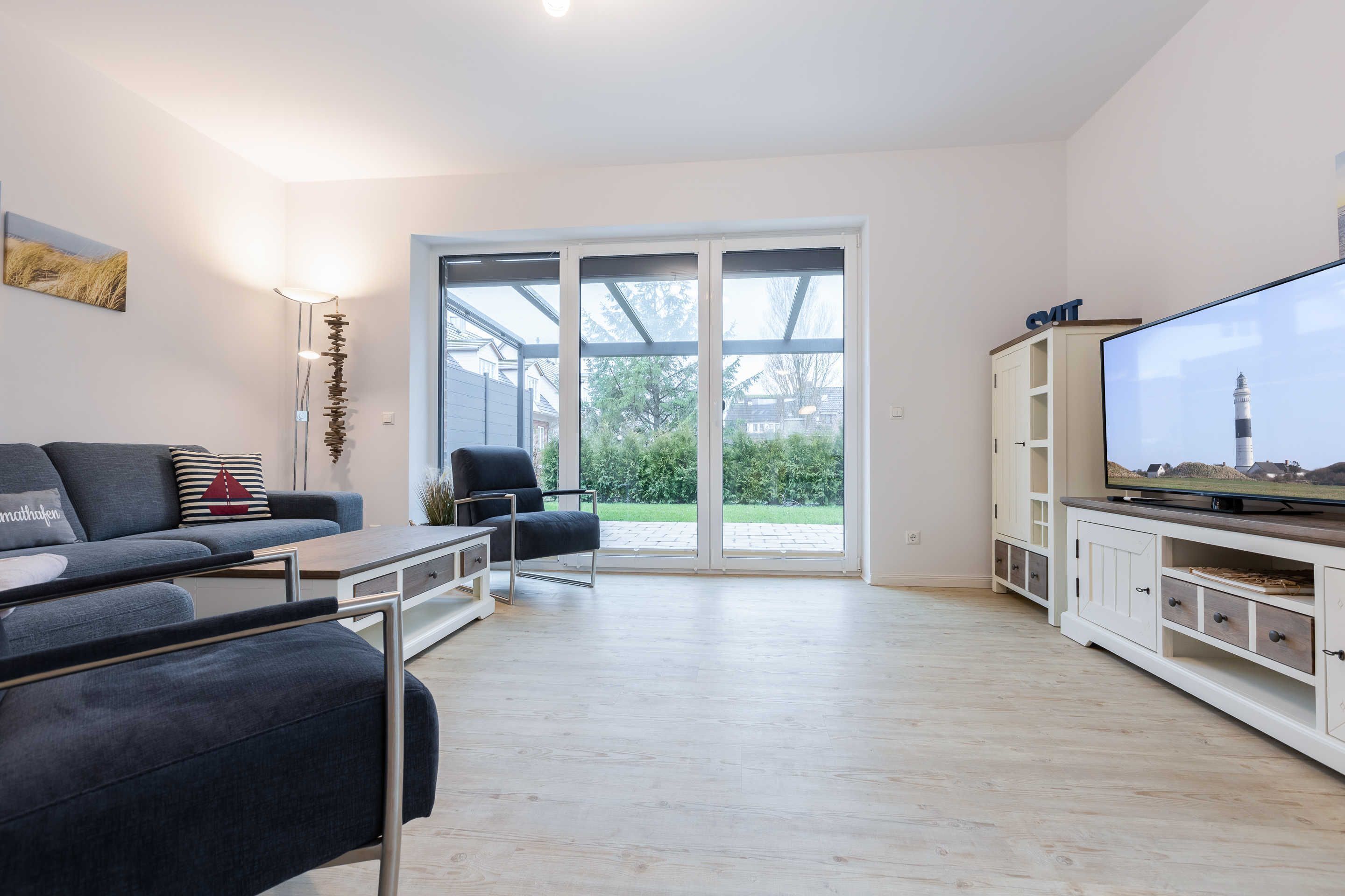 A modern living room with a sofa, armchair, TV stand, and a large window with a view of greenery.
