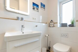 A bathroom with a white sink, mirror, and toilet. There are two wall-mounted light switches and a small window with a plant.