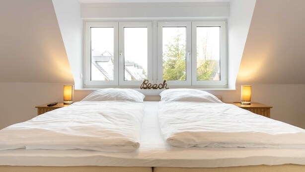 Un dormitorio con una cama grande, dos lámparas de noche y una ventana con vistas a edificios y árboles.