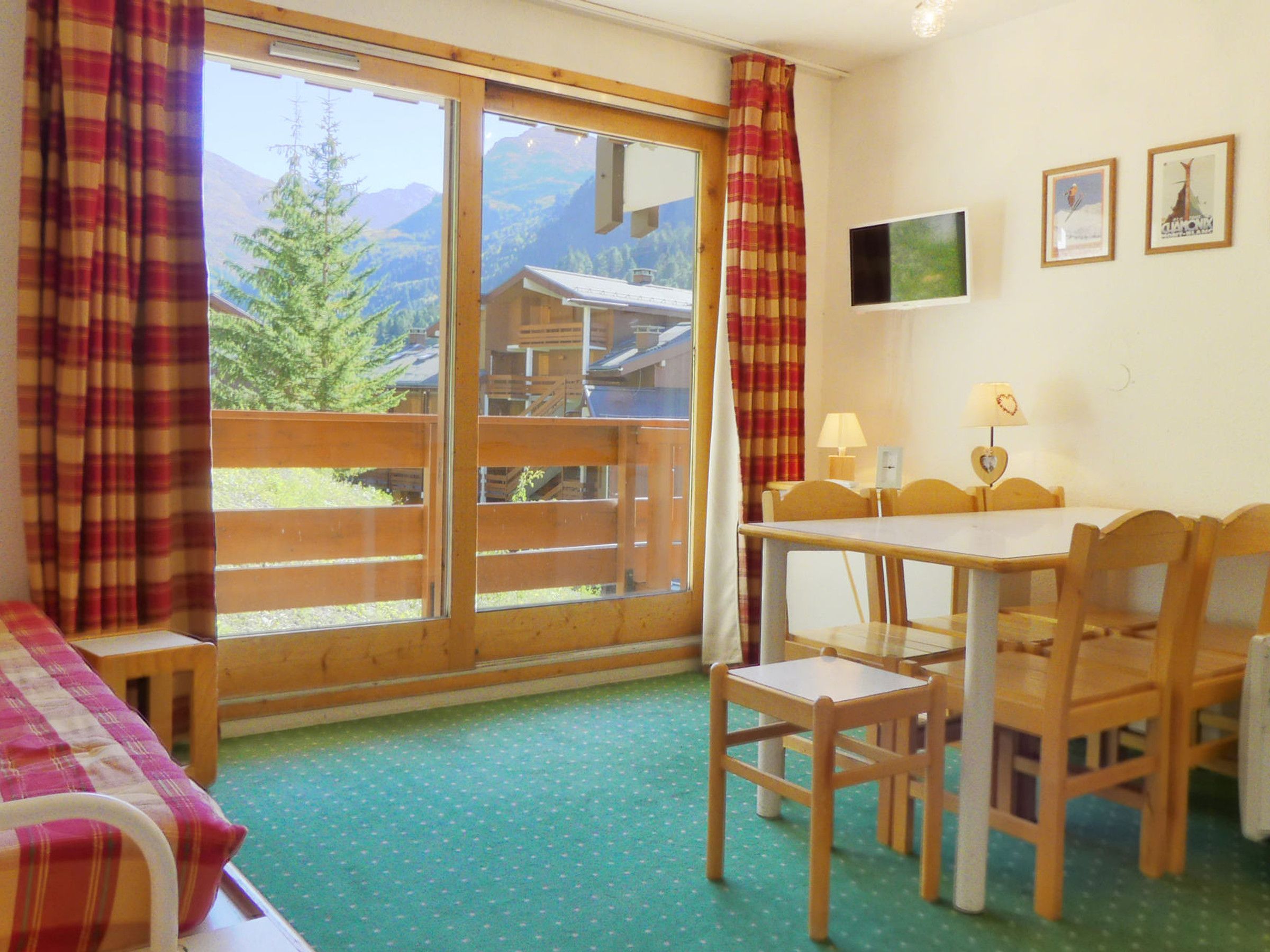 A room with a wooden table, chairs, a bed with plaid curtains, a TV, and a mountain view.