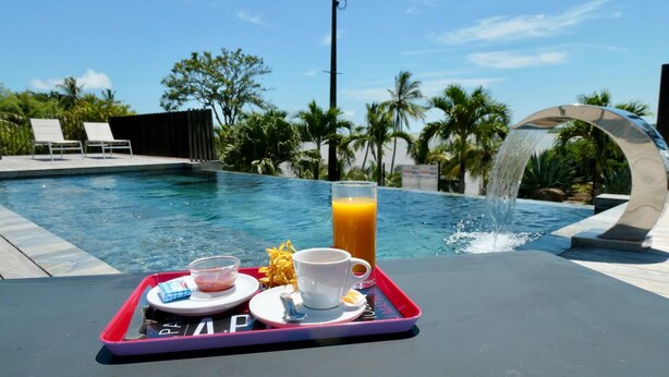 Un plateau de petit-déjeuner placé au bord de la piscine, avec un verre de jus d’orange, une tasse et un bol de nourriture, le tout sur fond de ciel bleu azur et de palmiers.