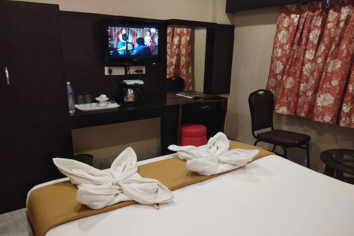 A hotel room with a bed, a TV on a stand, a desk with a chair, a red stool, and a window with floral curtains.