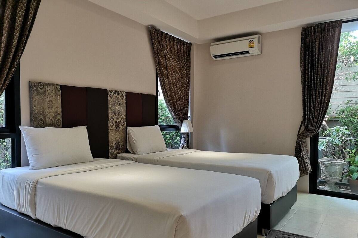 Hotelkamer met twee bedden, een raam met gordijnen, een airconditioning en een klein tafeltje met slippers.