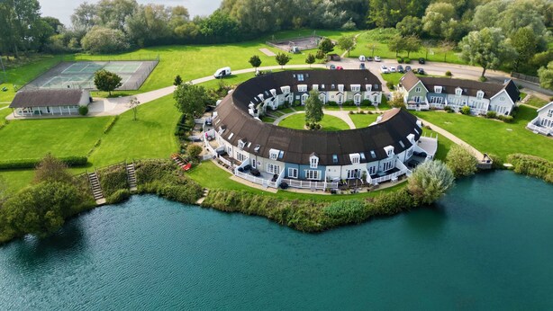 A circular building complex with a tennis court, surrounded by a lake and greenery.