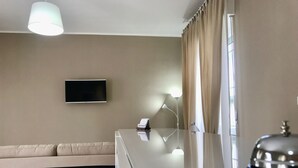 Habitación de hotel con televisor de pantalla plana en la pared, sofá, mesa de vidrio con teléfono y lámpara, y ventana con cortinas.