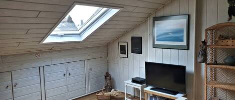 A room with a sloped ceiling, wooden cabinets, a television on a stand, a framed picture, and a wicker basket.