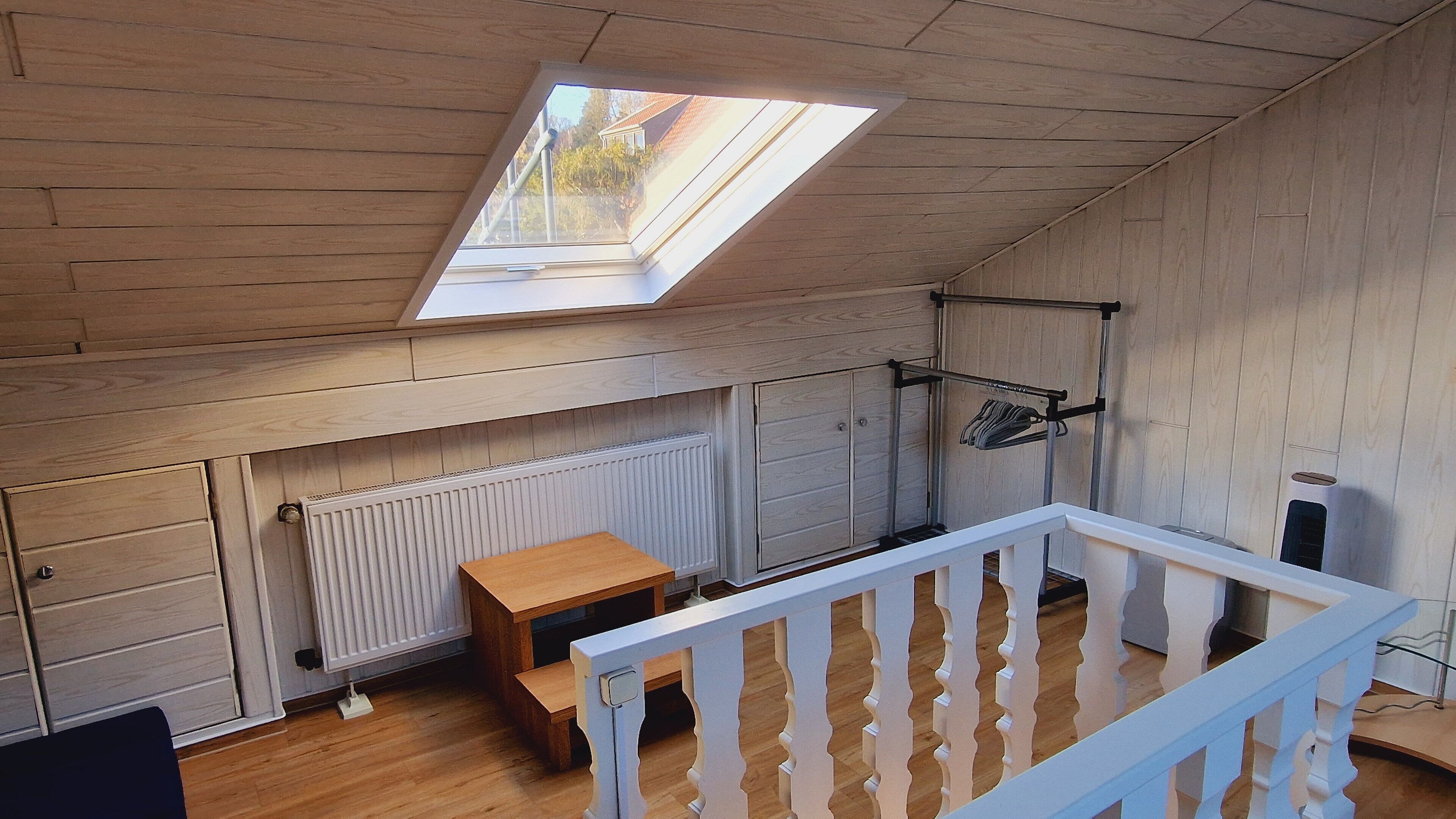 A room with a sloped ceiling, a skylight, a wooden table, a radiator, and a white railing.