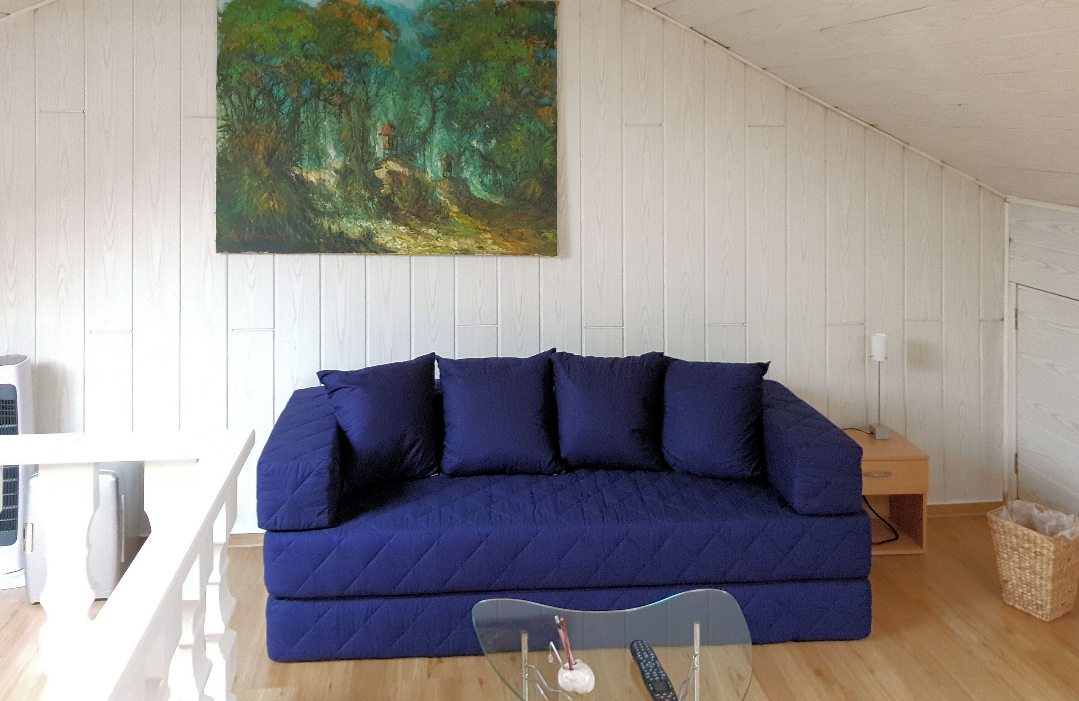Ein Zimmer mit einem dunkelblauen Sofa, einem Gemälde mit Waldszene, einer weißen Decke und Holzboden.