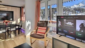A modern living room with a large window, a flat-screen TV, a dining area, and a cozy seating area.