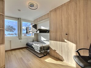 Quarto compacto com beliche, pequena escrivaninha, cadeira e janela com vista para uma paisagem nevada.