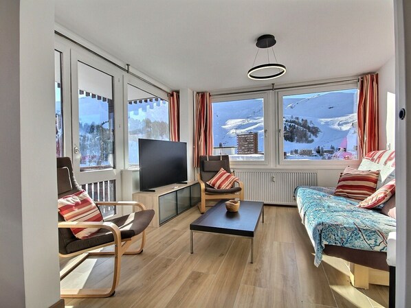 Uma sala de estar moderna com televisão, sofá e poltronas, grandes janelas com vista para uma montanha nevada.