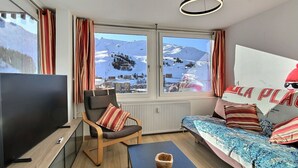 A modern room with a large window offering a mountain view, a sofa with striped cushions, a wooden chair, a flat-screen TV, and a small table with a bowl.