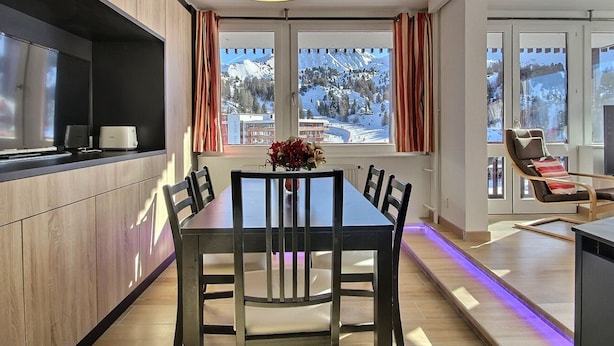 A modern dining area with a wooden table and chairs, a chandelier, and a view of a snowy mountain landscape through large windows.
