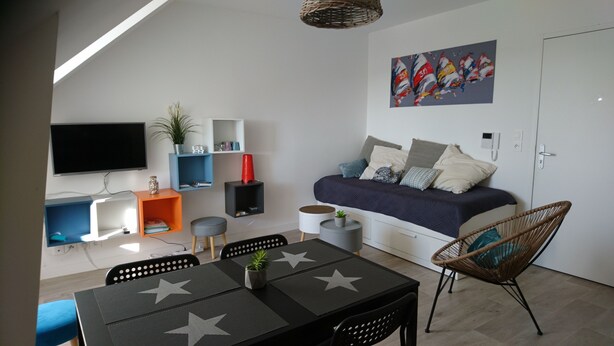 A modern, compact living space with a flat-screen TV, a bed with multiple pillows, a dining table with star-patterned placemats, and a wicker chair. - Emerald Coast Studio  (Saint-Méloir-des-Ondes)