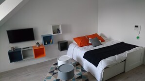 A modern bedroom with a sloped ceiling, a large bed with white bedding and orange pillows, a flat-screen TV mounted on the wall, and a geometric-patterned rug. - Emerald Coast Studio  (Saint-Méloir-des-Ondes)