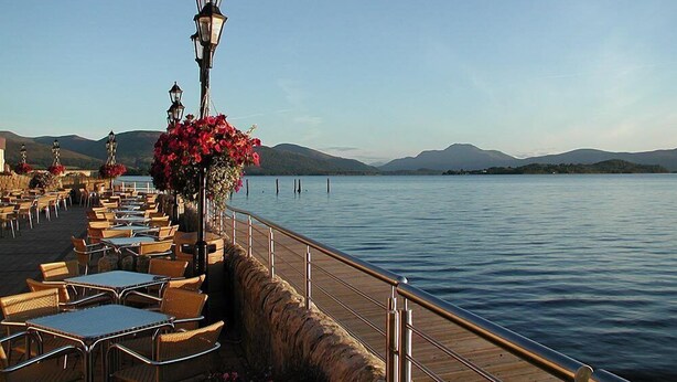 A waterfront dining area with outdoor seating, a stone wall, and a view of the lake and mountains. - Duck Bay Hotel (Alexandria)
