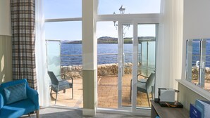 A room with a large sliding glass door leading to a patio with a view of the sea and hills. There are two chairs and a table on the patio. - Duck Bay Hotel (Alexandria)