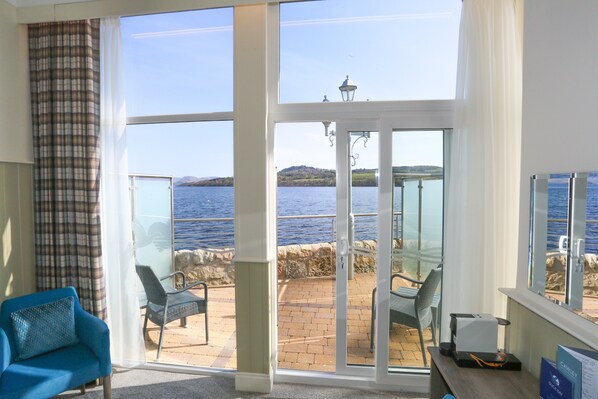 A room with a large sliding glass door leading to a patio with a view of the sea and hills. There are two chairs and a table on the patio.