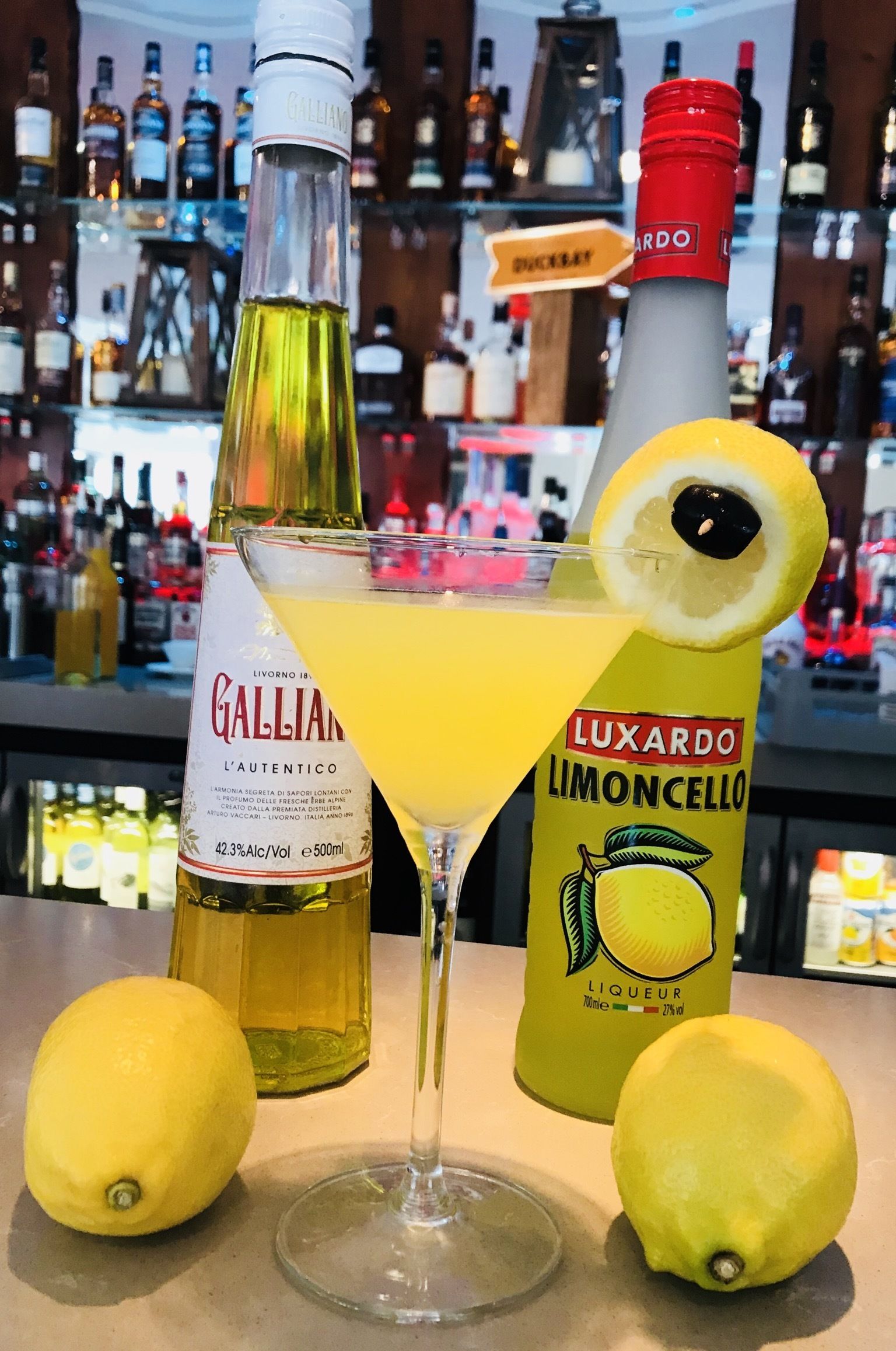 Un cóctel con una rodaja de limón, una botella de Galliano y una botella de Luxardo Limoncello.