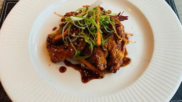 A white plate with a dish of food, possibly chicken, garnished with greens and sesame seeds, on a textured tablecloth.