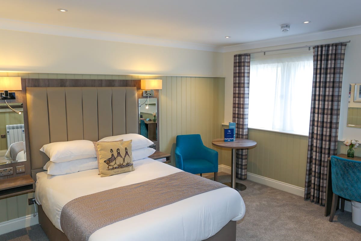 A hotel room with a large bed, a desk, a chair, a small table, and a window with curtains.