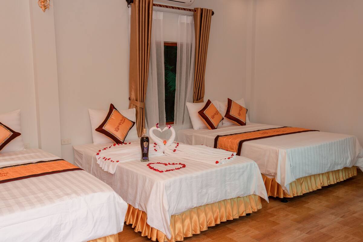 Une chambre d’hôtel avec trois lits, chacun recouvert d’une courtepointe blanche agrémentée de touches orange. Sur le lit central, on trouve une composition décorative comprenant une bouteille en forme de cœur et des pétales de rose.