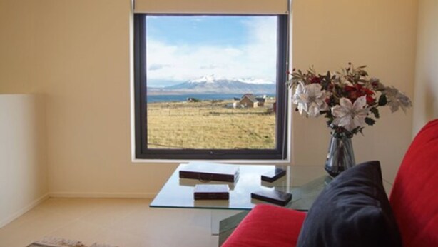 Una habitación con un ventanal que ofrece vistas a un campo y montañas. Hay una mesa de cristal con libros y un jarrón con flores.