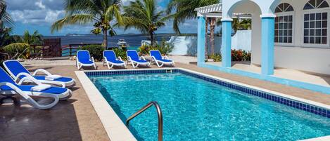 A pool with clear blue water, surrounded by blue lounge chairs and palm trees, with a view of the ocean and a building with arched windows.