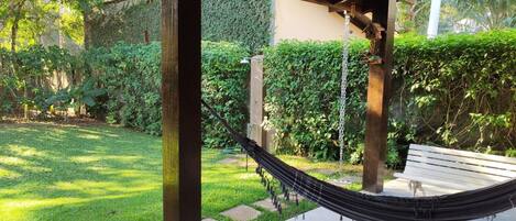 Una hamaca bajo una pérgola de madera en un jardín con un banco y abundante vegetación.