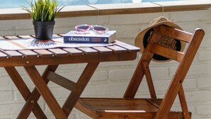 Ein Holztisch mit einem Blumentopf, einem Buch mit dem Titel 'Obsession' und einem Strohhut auf einem Balkon mit Blick auf das Meer.