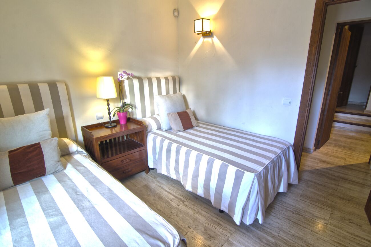 A hotel room with two beds, a nightstand with a lamp and a potted plant, and a wall-mounted light fixture.
