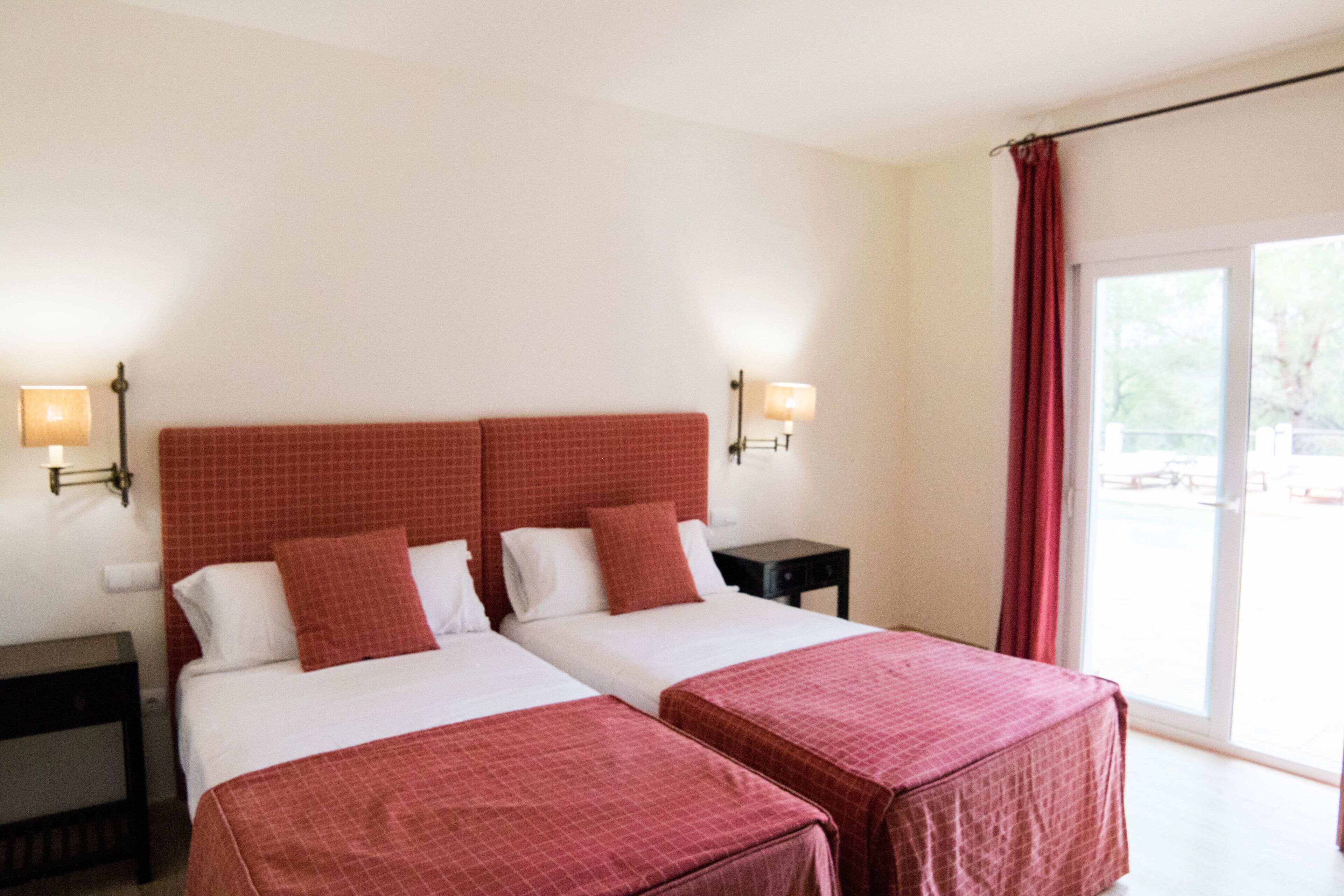 Ein Hotelzimmer mit zwei Betten, einem Nachttisch, einer Lampe und Blick ins Freie durch eine Schiebetür.