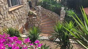Un bâtiment en pierre, doté d’un escalier en pierre menant à une porte, le tout entouré d’une végétation luxuriante et de fleurs roses éclatantes.