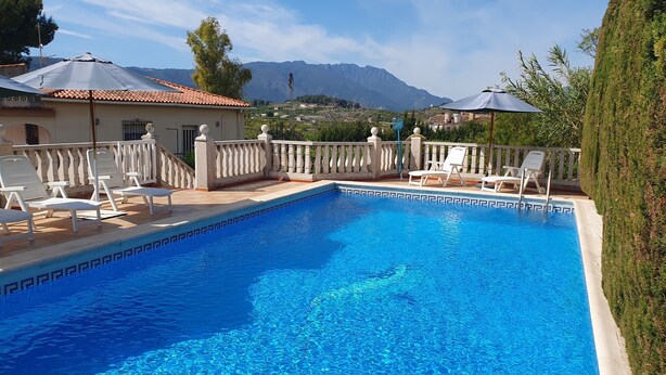 Une piscine dont le bord est revêtu de carrelage, entourée de chaises longues et de parasols, avec des montagnes en arrière-plan.