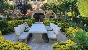 Un espace patio avec une table en pierre et des bancs, entouré de haies et de plantes bien entretenues.