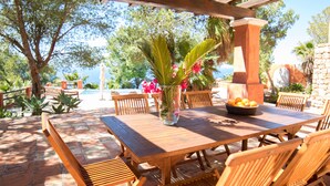 An outdoor wooden patio with a table, chairs, a vase with flowers, and a stone wall.
