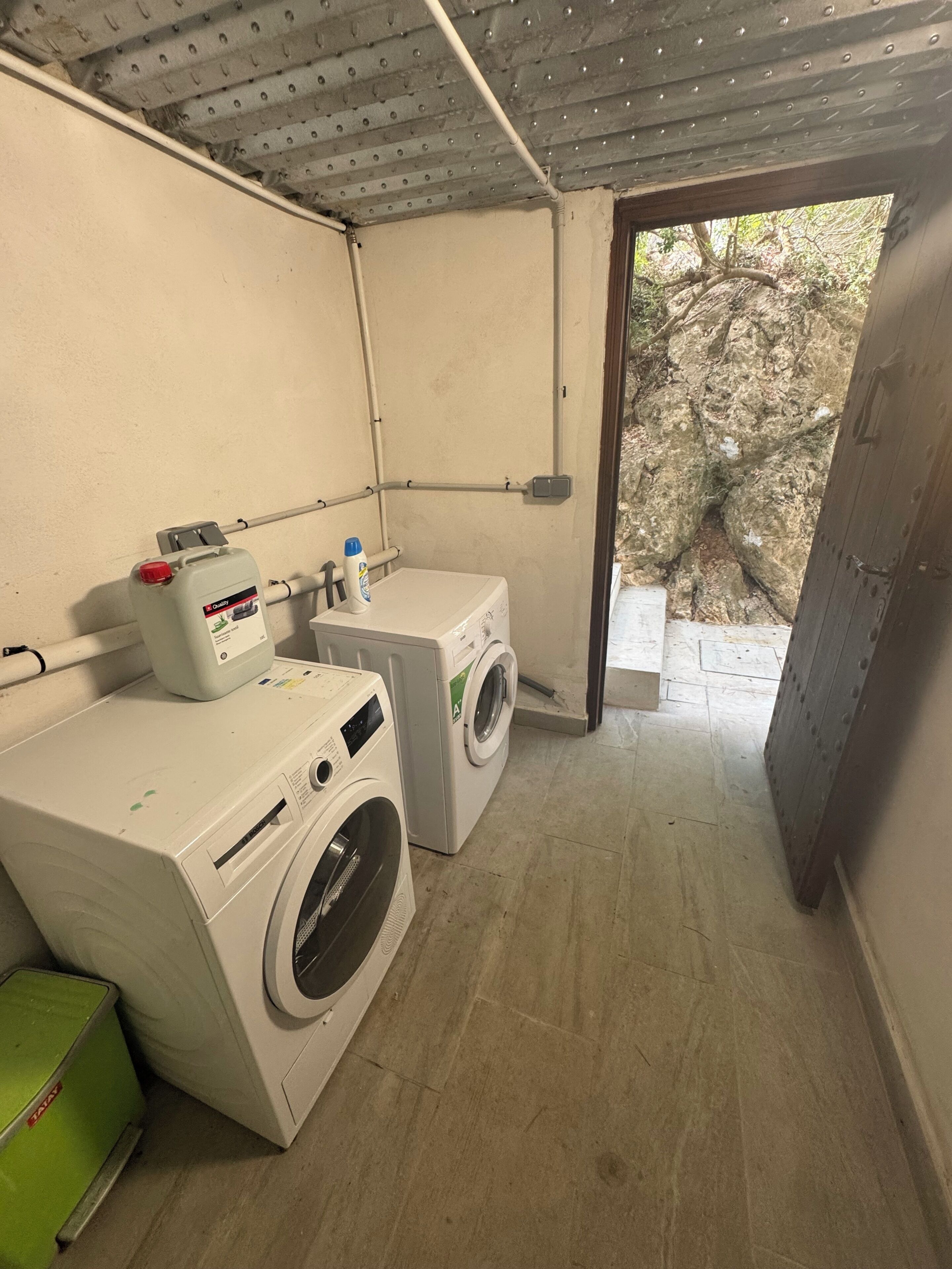 A laundry room with two washing machines, a detergent bottle, and a green trash bin.