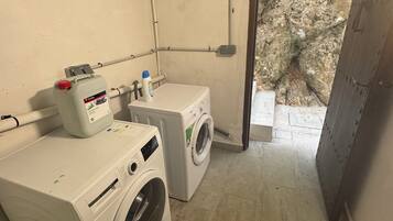 A laundry room with two washing machines, a detergent bottle, and a green trash bin.