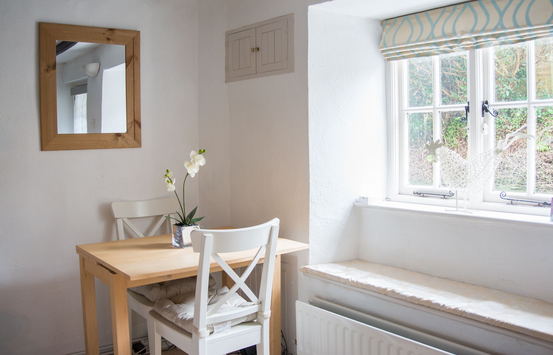 Un petit coin repas avec une table en bois et des chaises blanches, un miroir au mur et une fenêtre donnant sur un paysage verdoyant.
