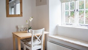 A small dining area with a wooden table and white chairs, a mirror on the wall, and a window with a view of greenery. - Pearl River Cottage in South Devon (Kingsbridge)