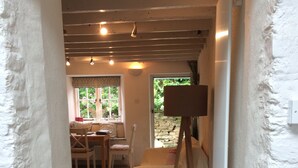 A narrow hallway with a wooden floor, a painting on a stand, a dining area with a table and chairs, and a window with a view of greenery. - Pearl River Cottage in South Devon (Kingsbridge)