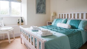 A bedroom with a bed, a chair, a nightstand, a lamp, and a window with curtains. - Pearl River Cottage in South Devon (Kingsbridge)